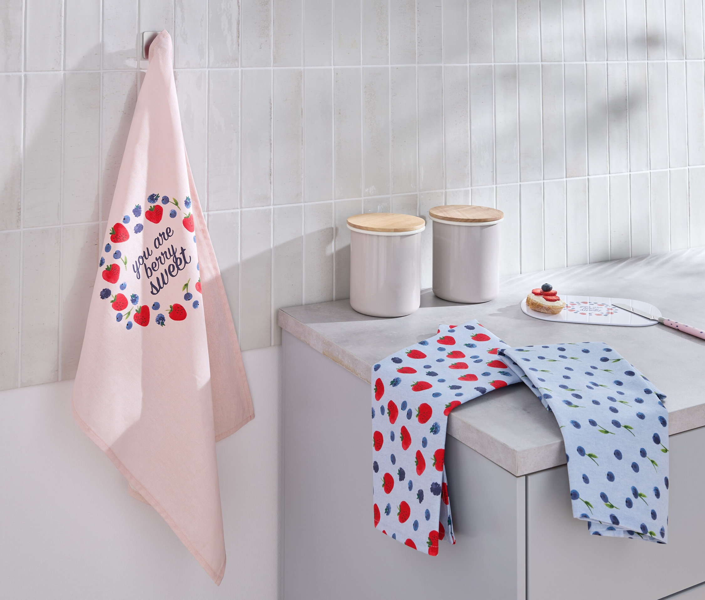 Egy világos rózsaszín, eper- és áfonyamintás konyharuha lóg a falon. A munkalapon két összehajtott konyharuha és két fehér, fa fedéllel ellátott kanna található.