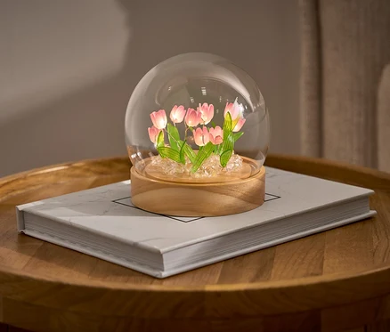 LED-DIY-Bastel-Set tulipánokkal egy üvegkupolában egy könyvön egy kerek asztalon.