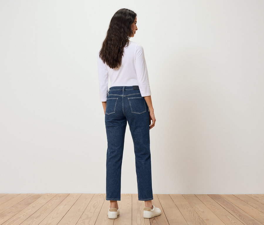 Egy sötét hajú nő áll háttal a kamerának, kék farmerban és fehér pólóban.