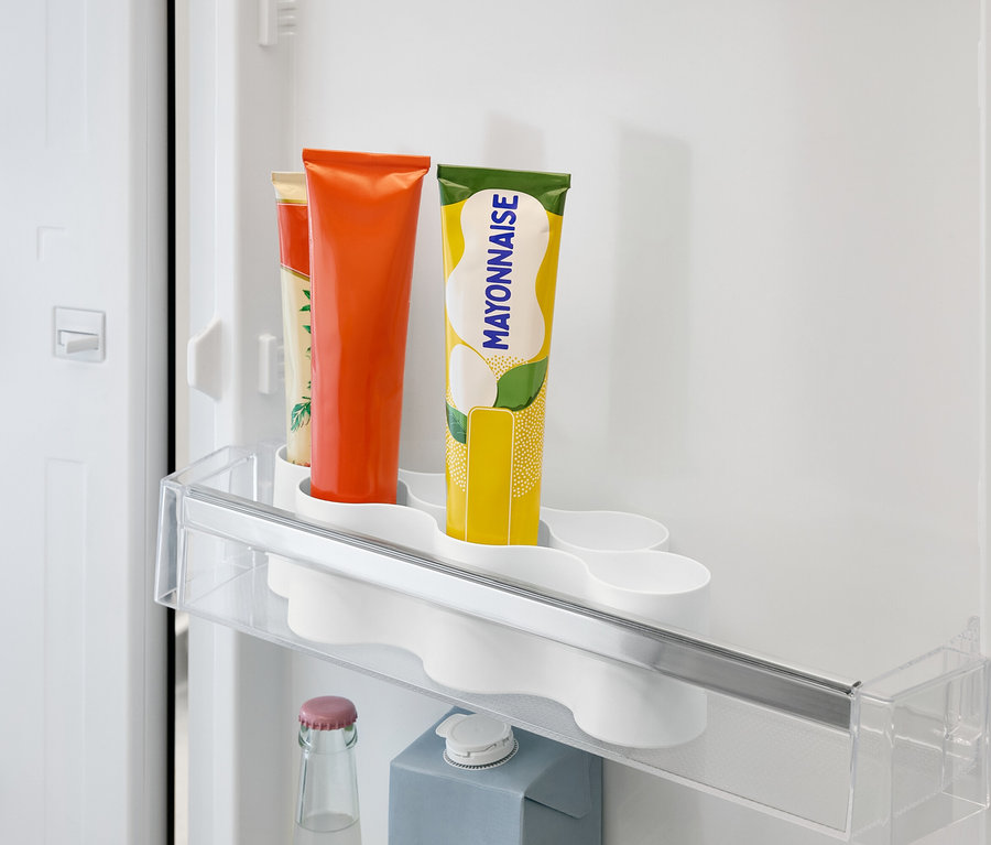 A hűtőszekrényben van egy átlátszó polc narancssárga, sárga és bézs címkés tubusokkal, alatta pedig egy üveg és egy karton.