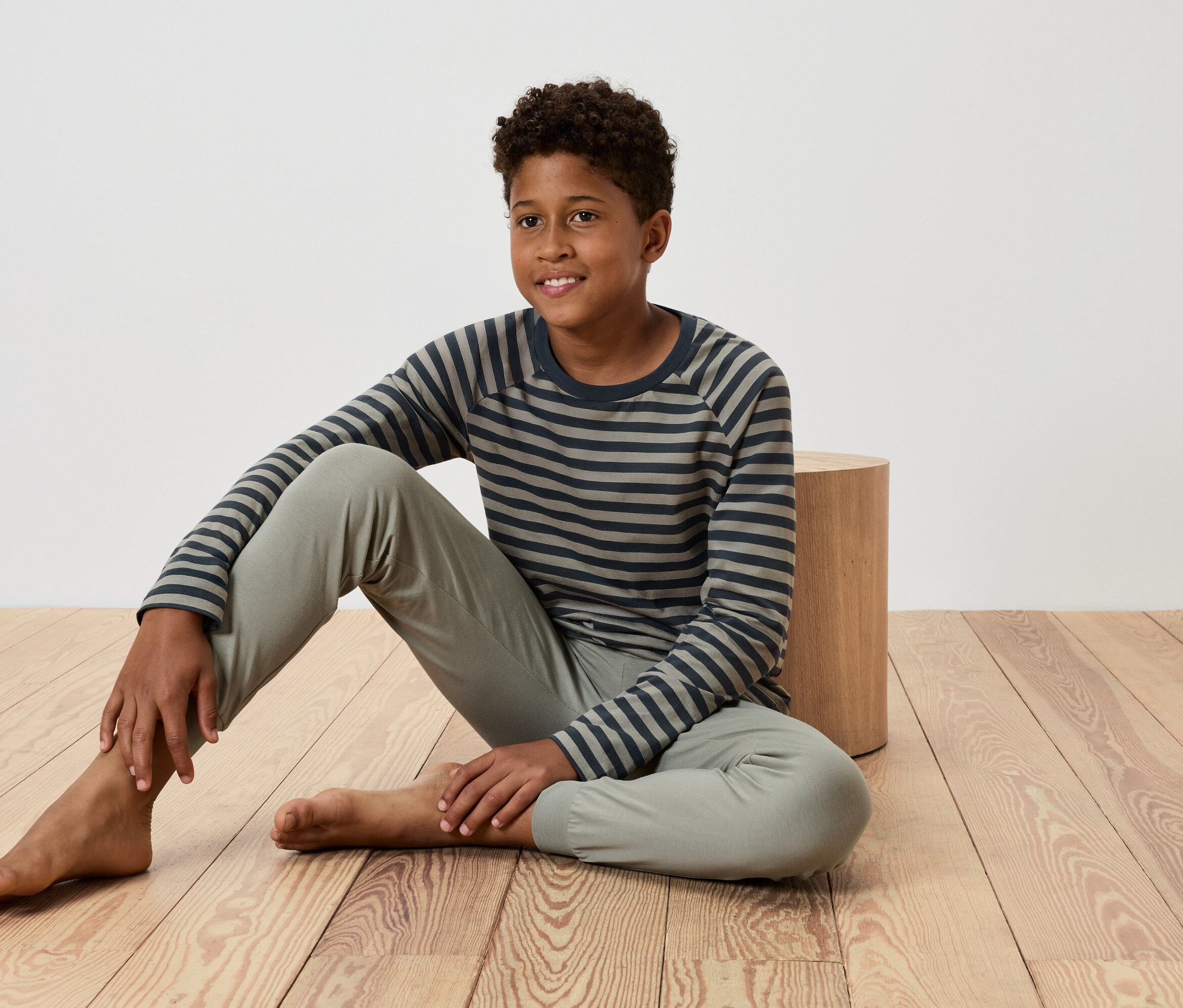 Egy fiatal fiú ül egy fapadlón, egy kis faasztalnak támaszkodva, csíkos pólót és nadrágot visel.