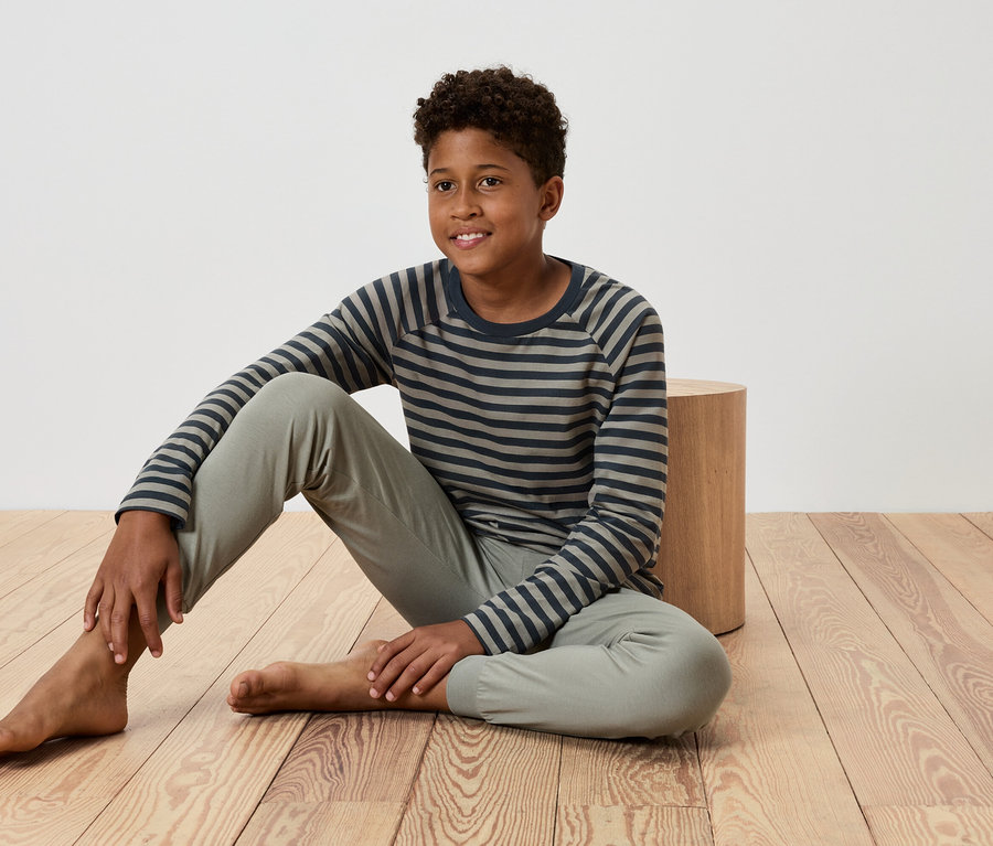 Egy fiatal fiú ül egy fapadlón, egy kis faasztalnak támaszkodva, csíkos pólót és nadrágot visel.