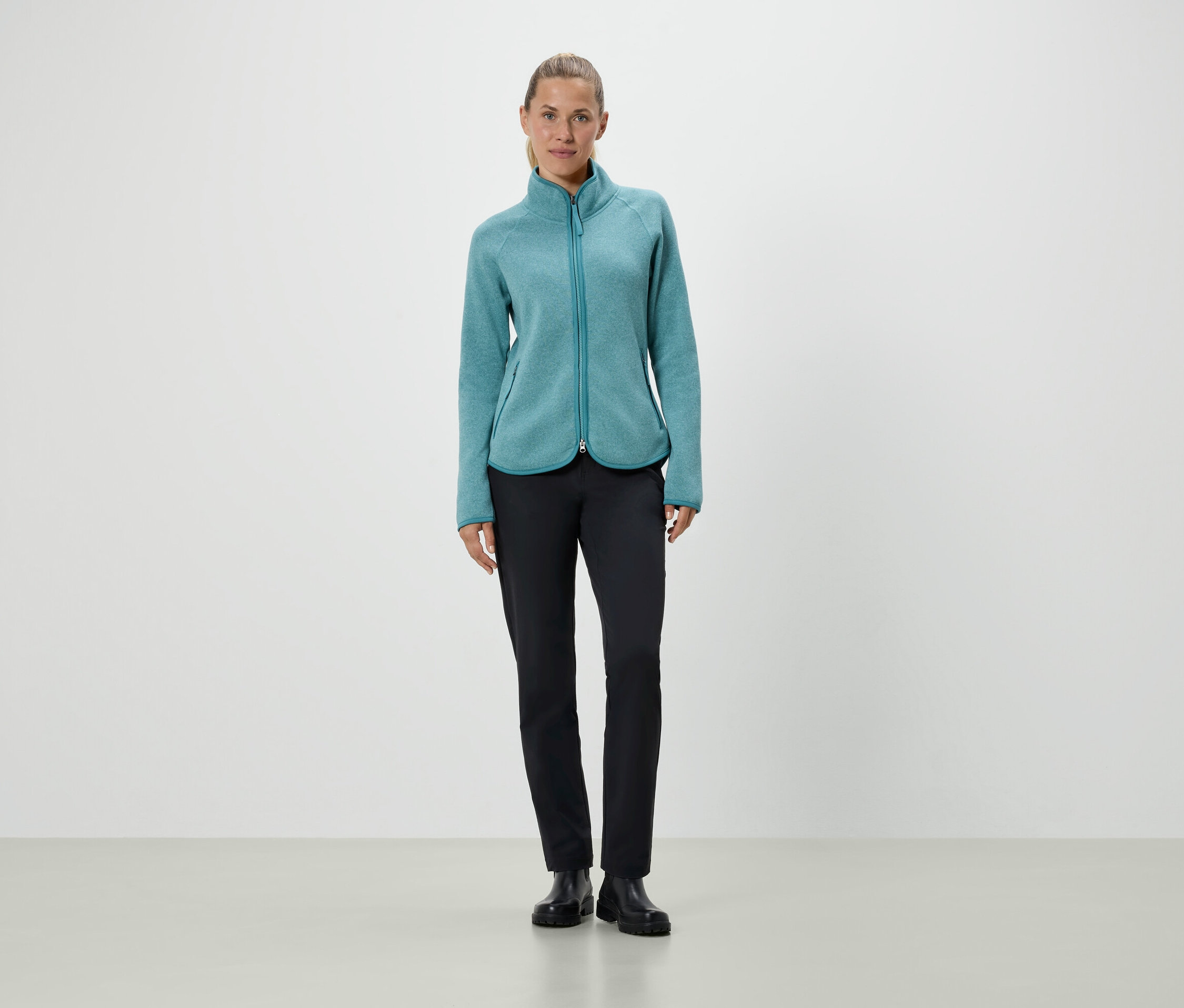 Nő áll türkiz színű fleece kabátban és fekete nadrágban.
