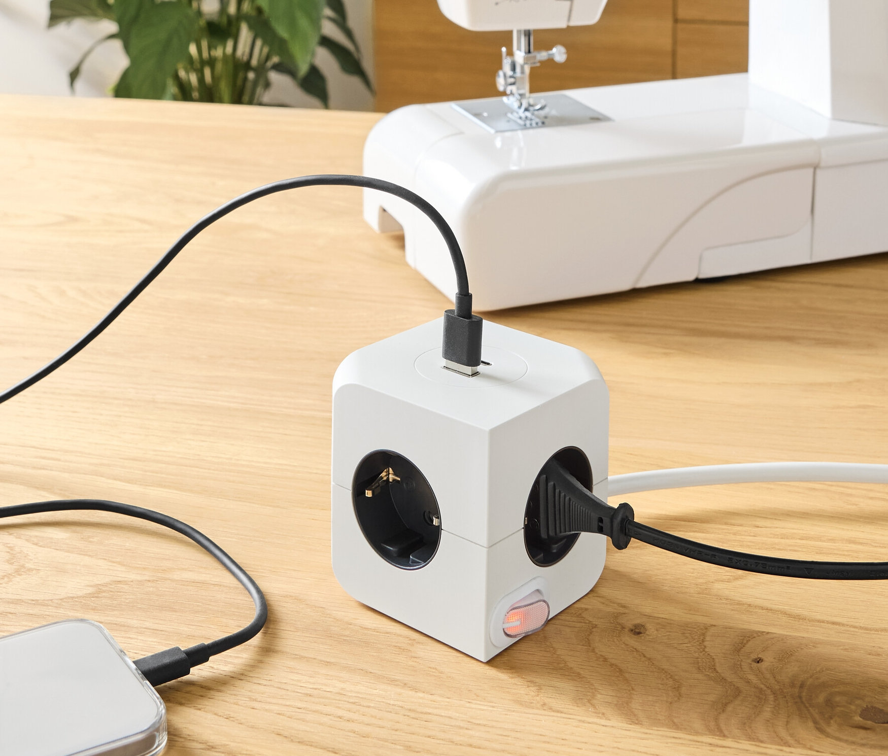 Egy fa asztalon egy USB-s dugaszoló kocka van, amihez egy telefon és egy varrógép van csatlakoztatva.