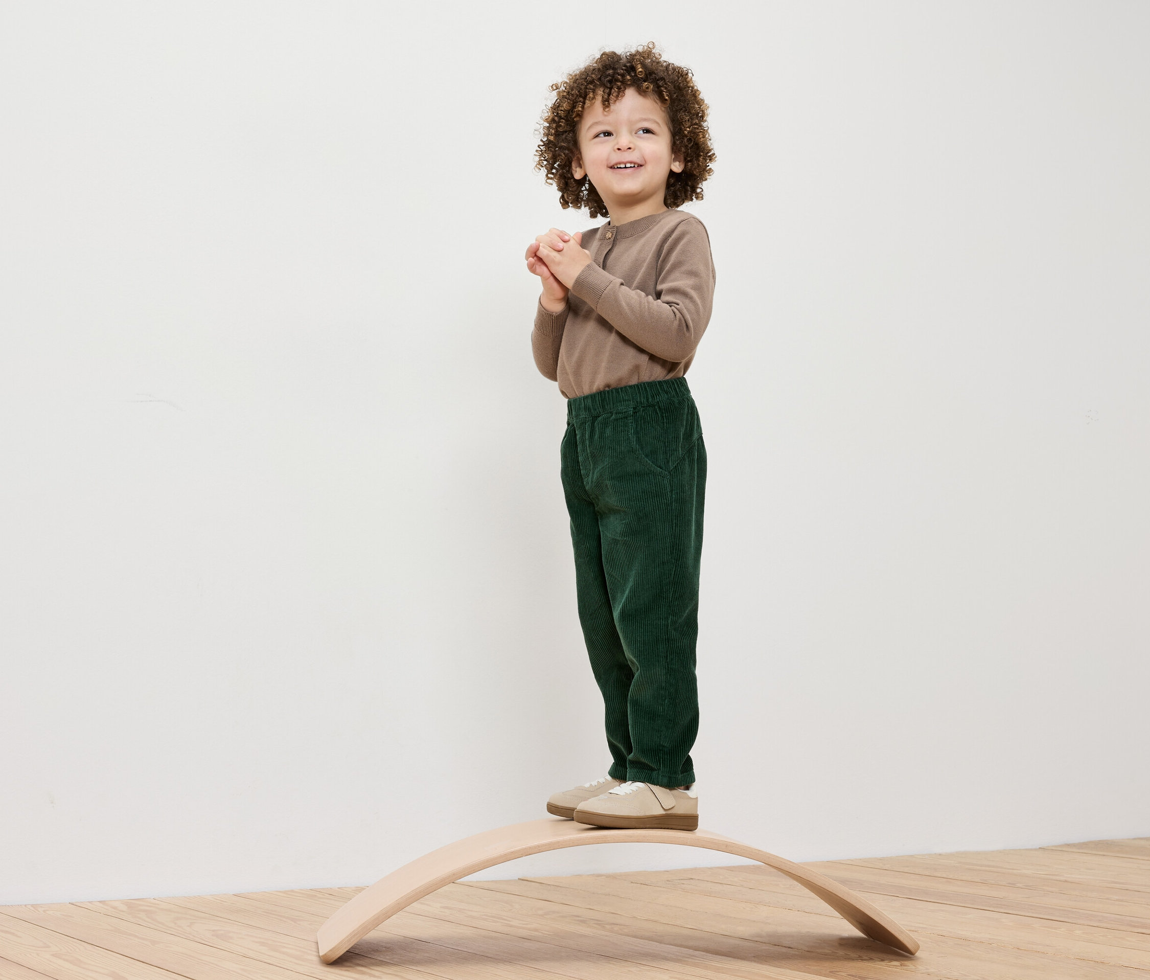 Egy göndör hajú fiú áll egy fa egyensúlyozó deszkán, barna inget és zöld nadrágot visel.