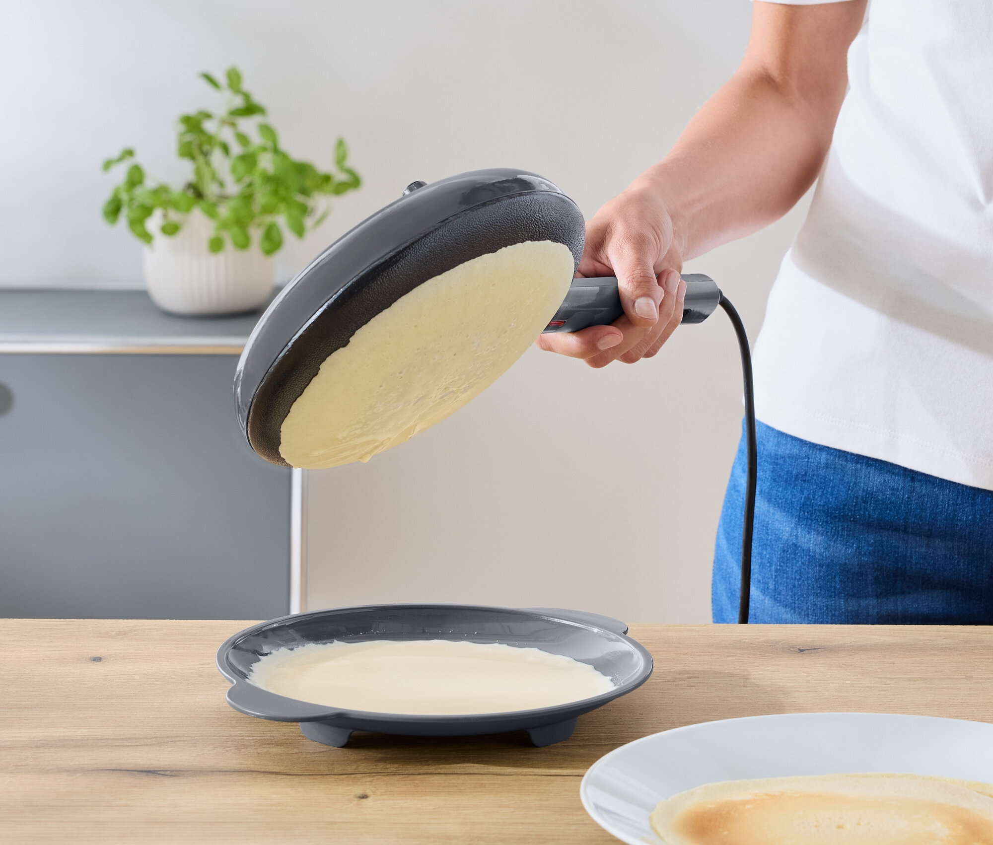 Egy személy egy sötétszürke elektromos serpenyőt egy tésztamaradékot tartalmazó tányér fölé dönt. Egy kész palacsinta egy fehér tányéron fekszik egy fa pulton.