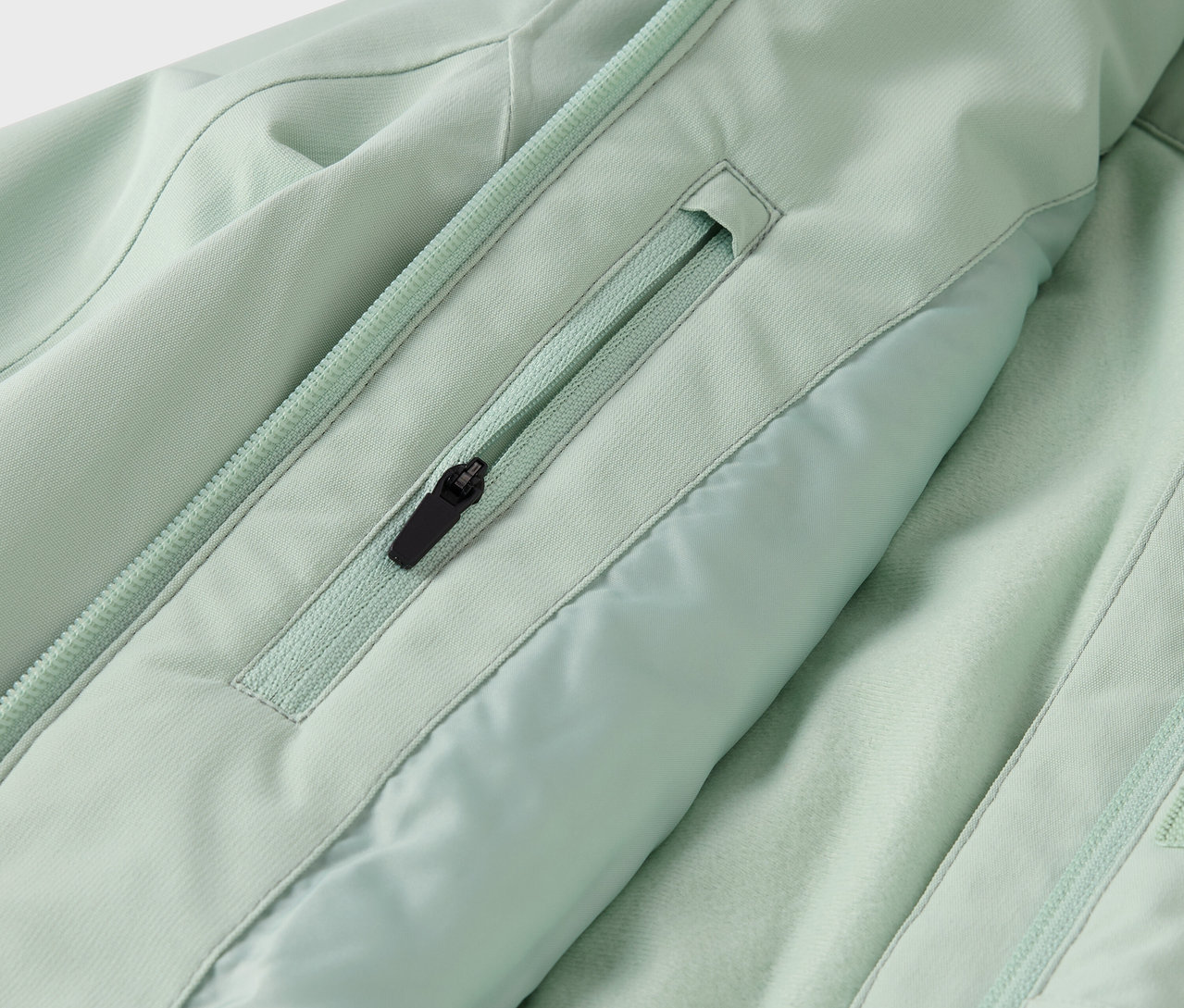 Egy világoszöld kabát részlete egy cipzáras zsebbel és egy cipzárral a közepén.