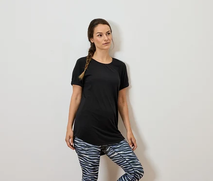 Barna hajú nő fekete pólóban és mintás leggingsben áll egy fehér falnál.