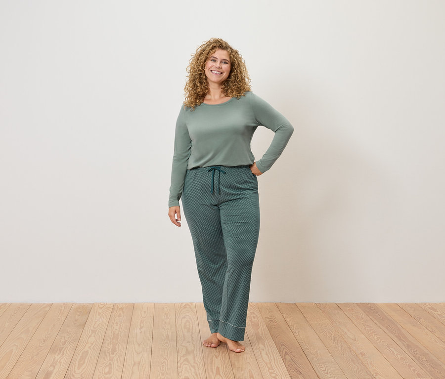 Nő áll és pózol zöld pizsamában egy fa padlón egy fehér fal előtt.