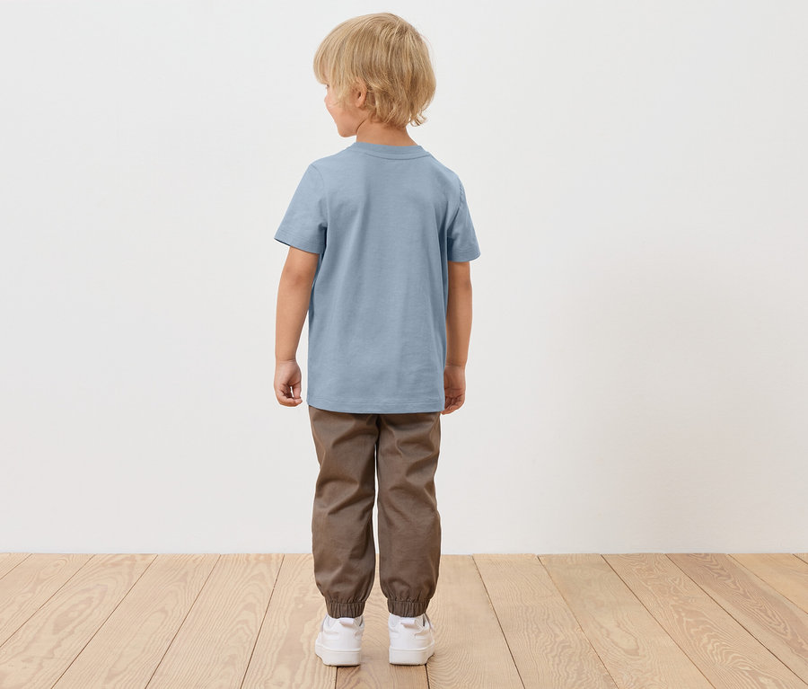 Egy szőke hajú fiú háttal áll, kék pólót, barna nadrágot és fehér cipőt visel.