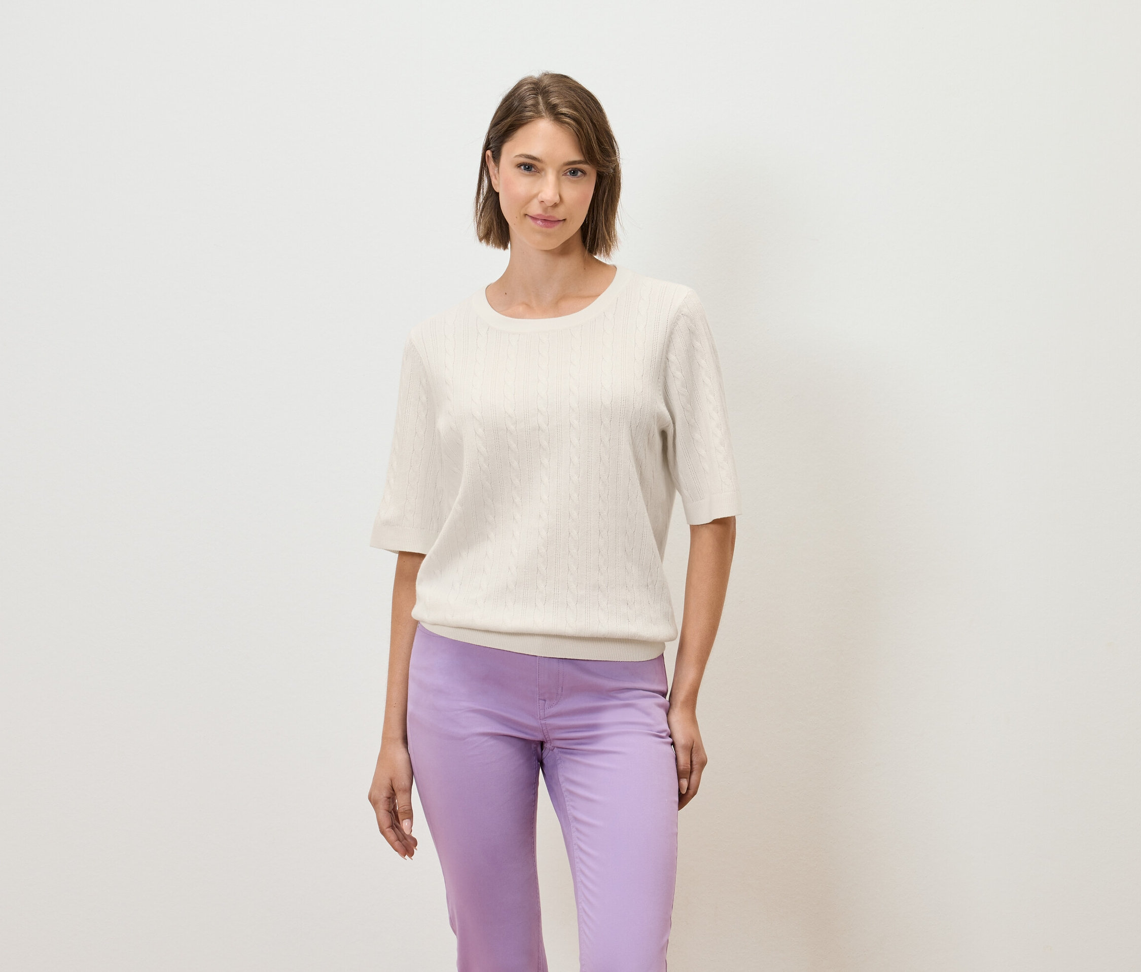 Egy nő világos háttér előtt pózol krémszínű, rövid ujjú pulóverben és lila nadrágban.