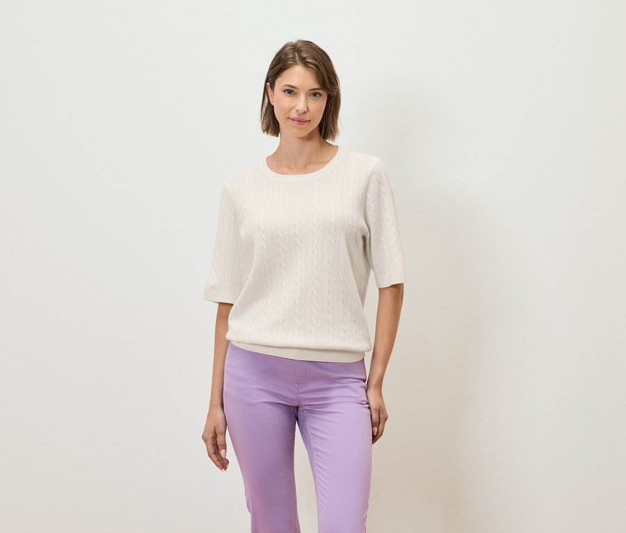 Egy nő világos háttér előtt pózol krémszínű, rövid ujjú pulóverben és lila nadrágban.