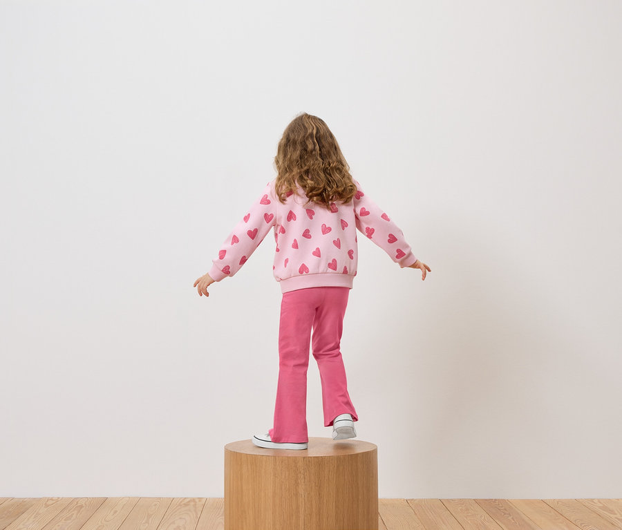 Egy lány háttal áll a kamerának egy faemelvényen, rózsaszín, szívecskés pulóvert és rózsaszín nadrágot visel.