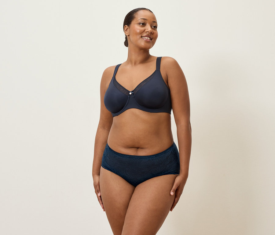 Egy középkorú, sötét hajú, tengerészkék alsóneműt viselő nő áll egy krémszínű háttér előtt.