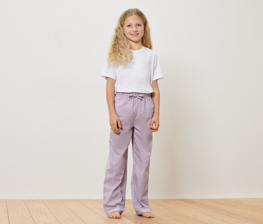 Egy szőke, göndör hajú lány fehér pólóban és lila csíkos nadrágban mezítláb áll egy fapadlón egy fehér fal előtt.