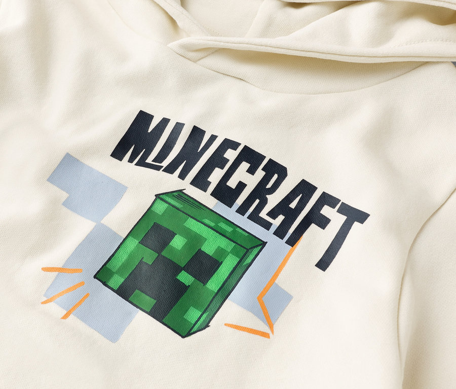 Egy krémszínű kapucnis pulóver részletfotója "Minecraft" nyomattal és egy zöld Creeper kockával.