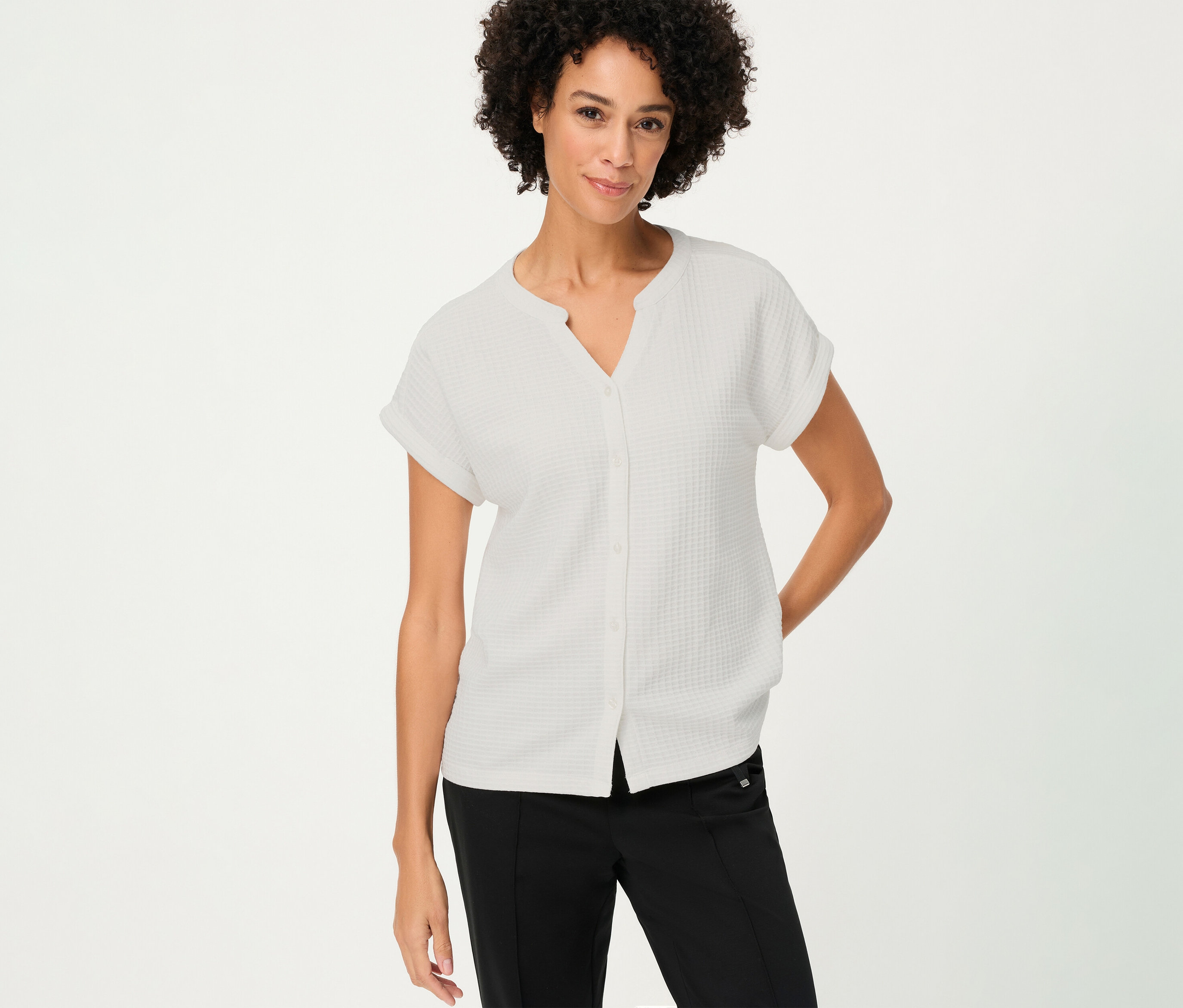A göndör hajú nő egy fehér, strukturál Olsen pólót visel gombokkal és egy fekete nadrágot.