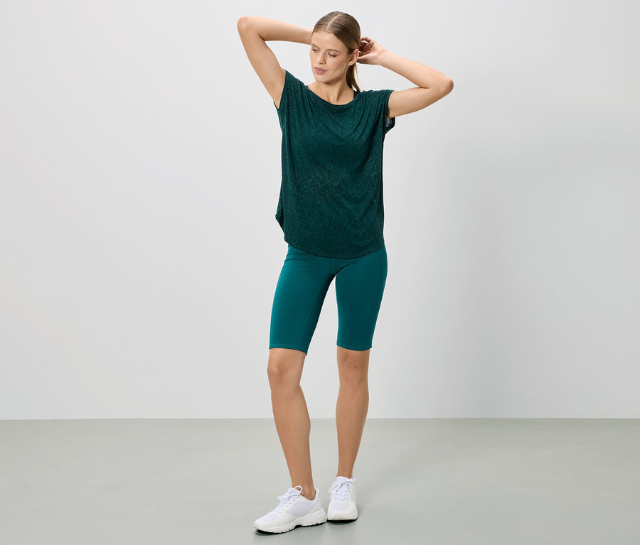 Nő pózol sötétzöld pólóban és rövidnadrágban, valamint fehér tornacipőben.