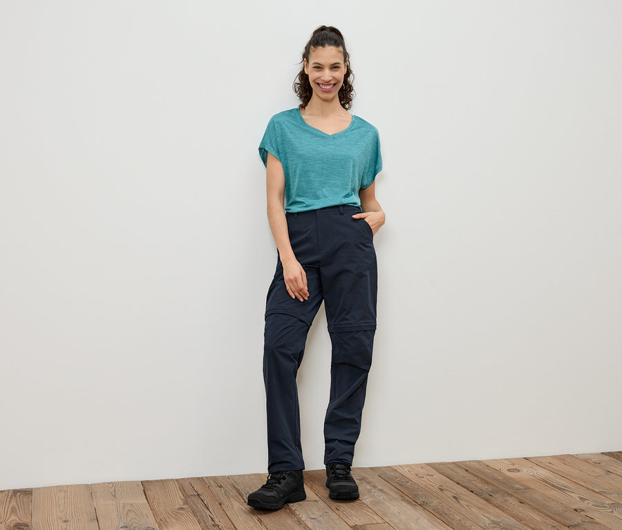 Egy nő áll egy fehér fal előtt, türkizkék pólót, sötétkék, lecipzárazható nadrágot és fekete cipőt visel.