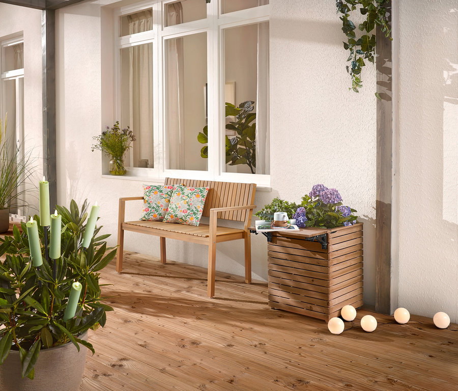 A fa teraszon egy fából készült pad párnákkal, egy virágláda virágokkal és egy cserepes növény gyertyákkal található.