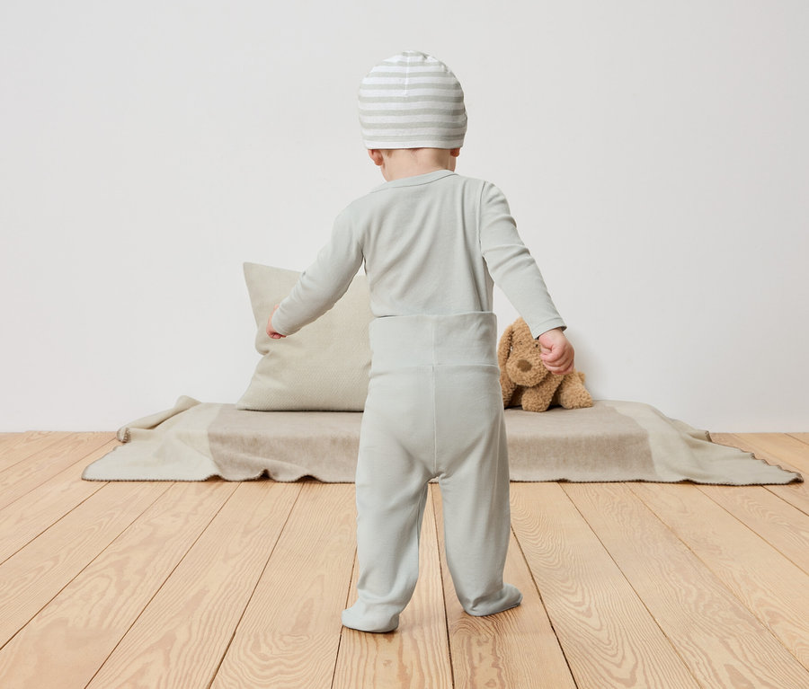 Egy kisgyermek világoszöld pizsamában és csíkos sapkában áll háttal a kamerának egy fapadlón.