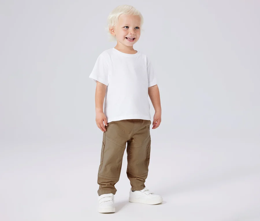 Mosolygó fiú fehér pólóban, khaki NAME IT twillnadrágban és fehér cipőben.