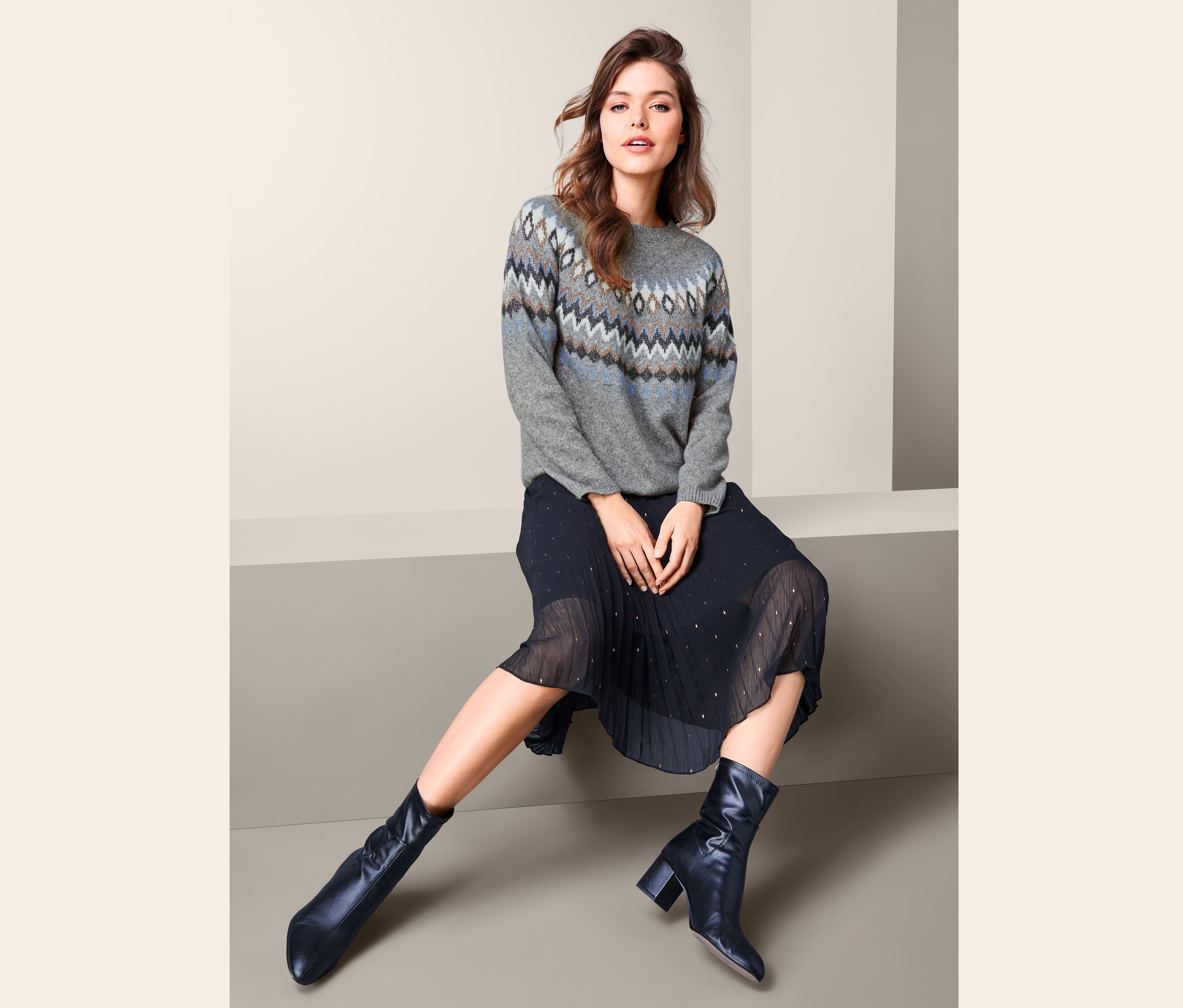 Nő ül, és norvégmintás kötött pulóvert, pliszírozott szoknyát és bőrcsizmát visel.