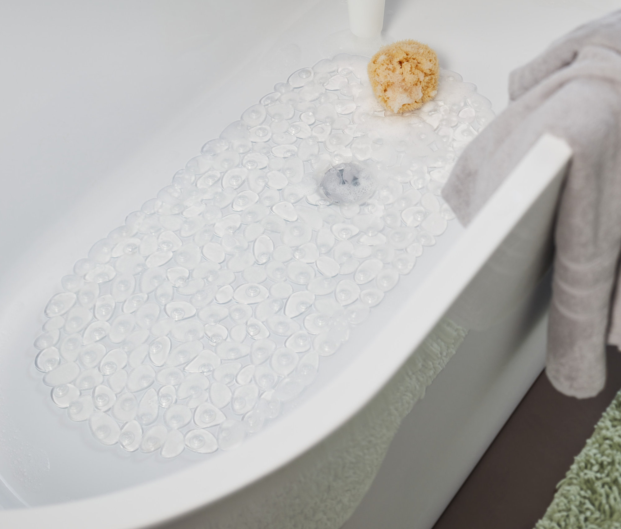 Egy átlátszó fürdőkádbetét van a fürdőkádban. A fürdőkád szélén egy szürke törölköző lóg, a fürdőkád mellett pedig egy zöld szőnyeg van.