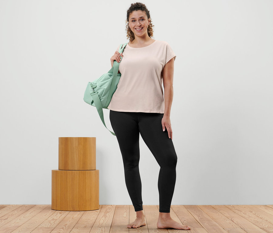 Nő pózol krémszínű sportfelsőben, fekete sportnadrágban és zöld sporttáskával a vállán.