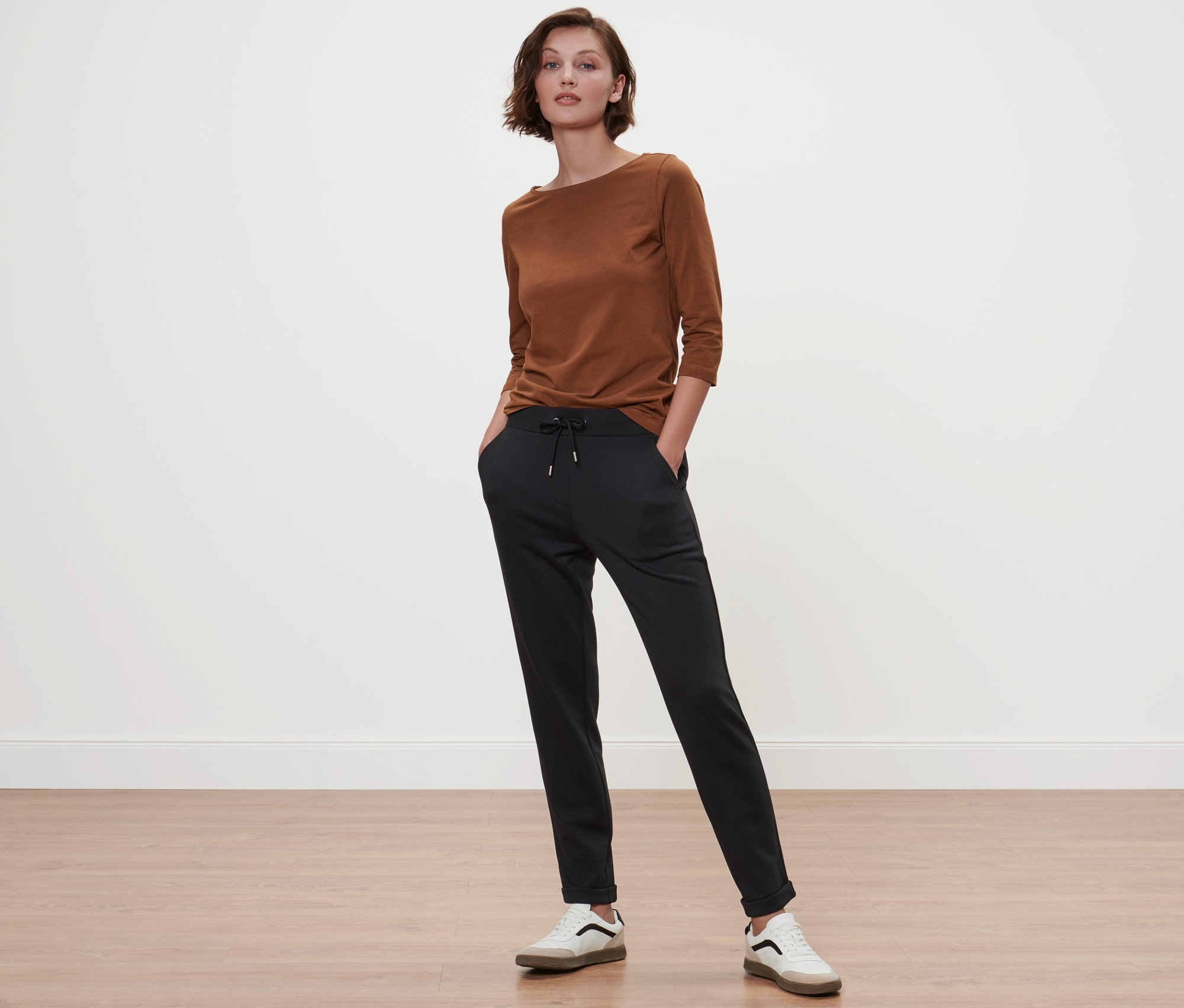 Nő áll fekete nadrág zsebében a kezével, és egy konyak színű, 3/4-es ujjú felsőt visel.