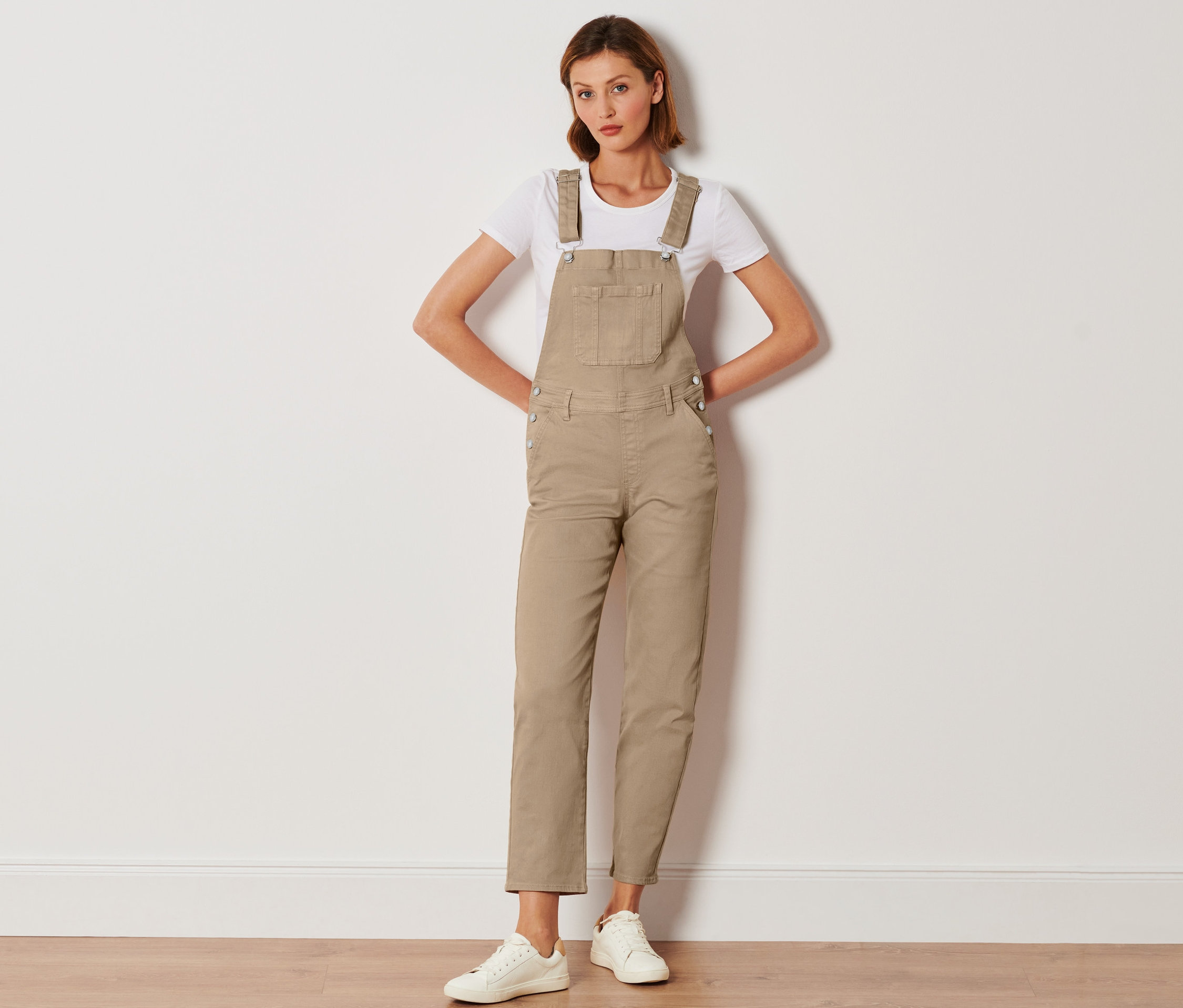 Nő pózol bézs női kantáros nadrágban, fehér pólóban és fehér tornacipőben.