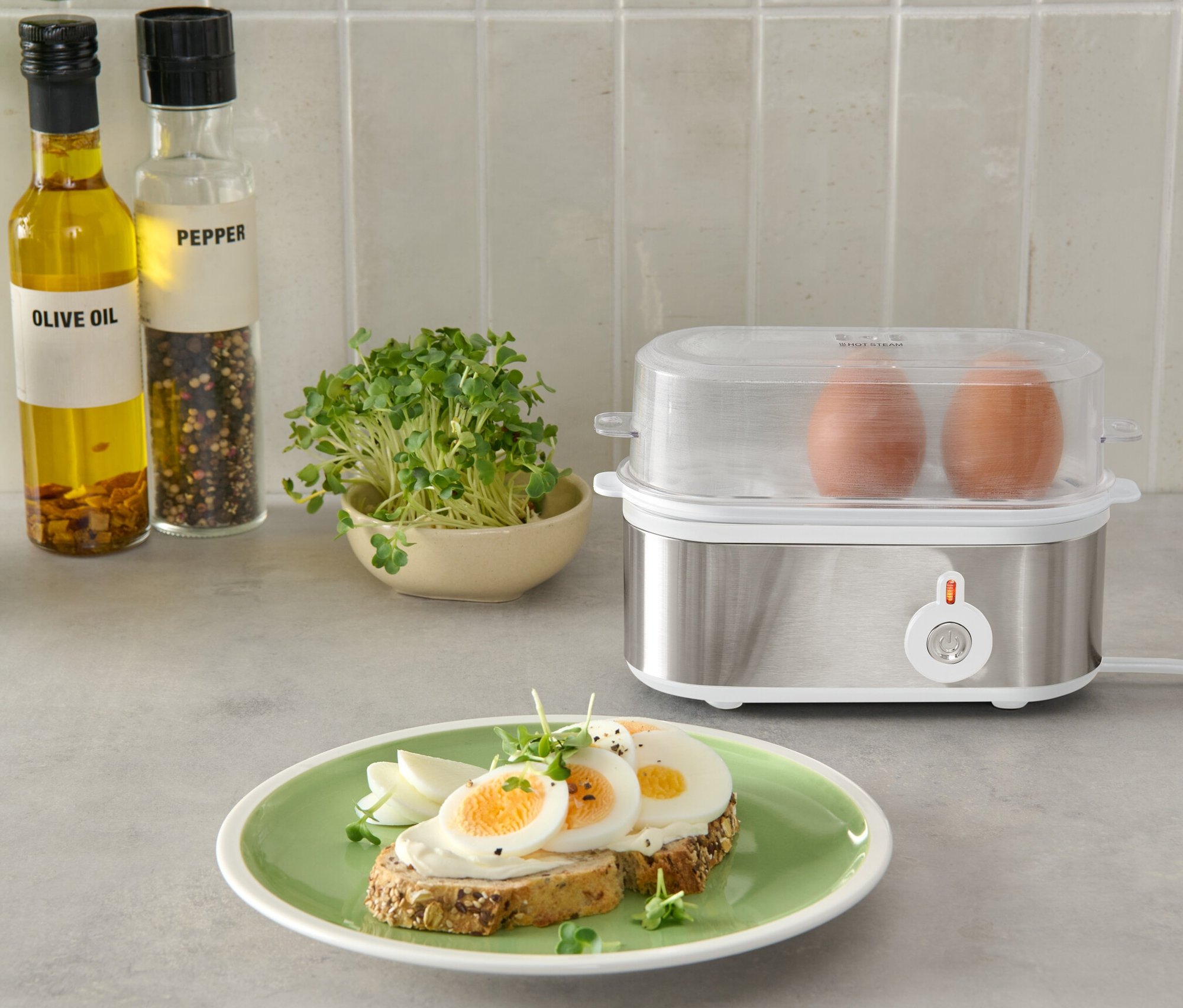 Két barna tojás egy ezüst tojásfőzőben, átlátszó fedővel, egy tányér kenyér és tojás mellett.