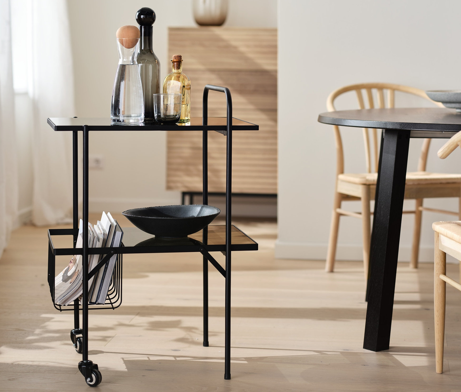 Fekete zsúrkocsi üvegpolcokkal és kerekekkel, italokkal és egy tálalóedénnyel.