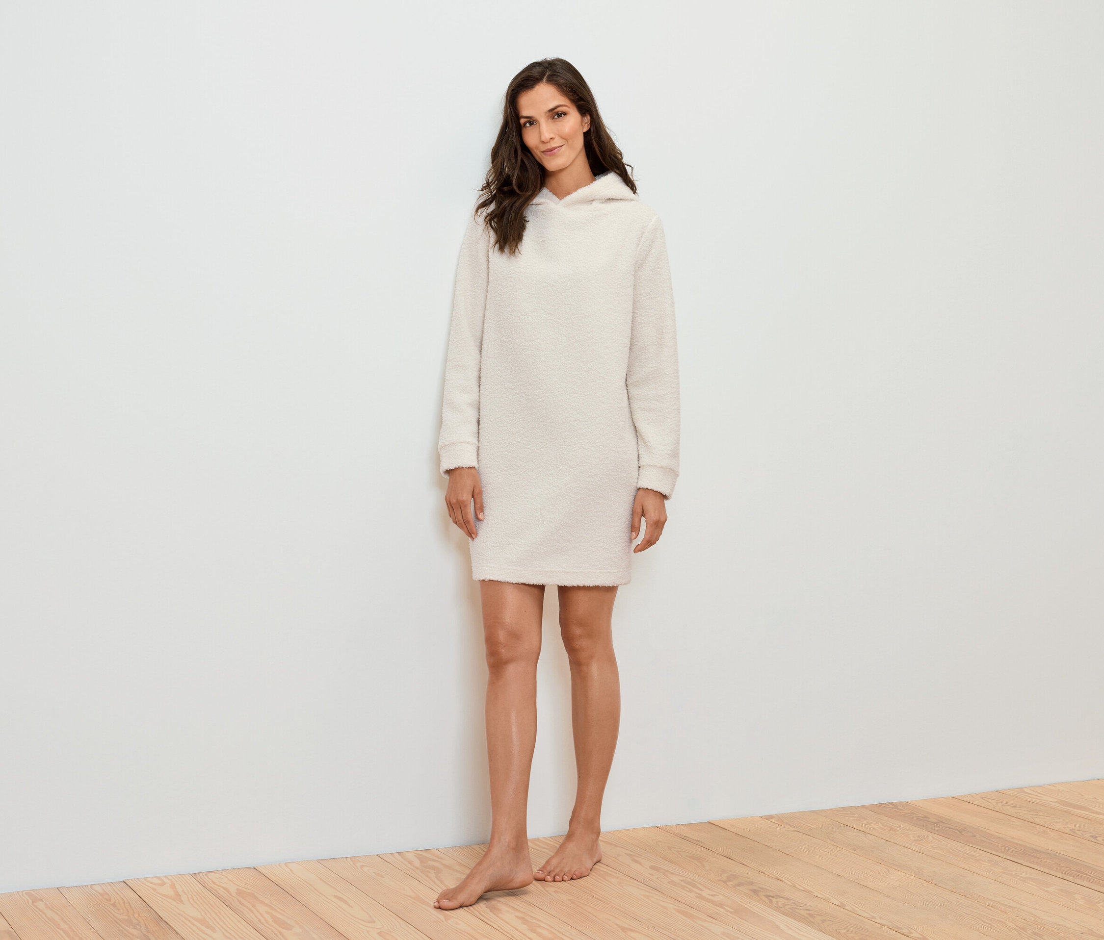 Egy nő áll egy fehér fal előtt, és egy krémszínű kapucnis ruhát visel.