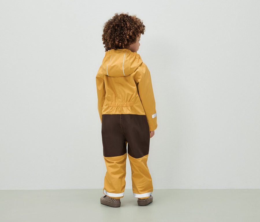  göndör hajú gyermek áll háttal a kamerának, barna-mustársárga, kapucnis overallt visel.