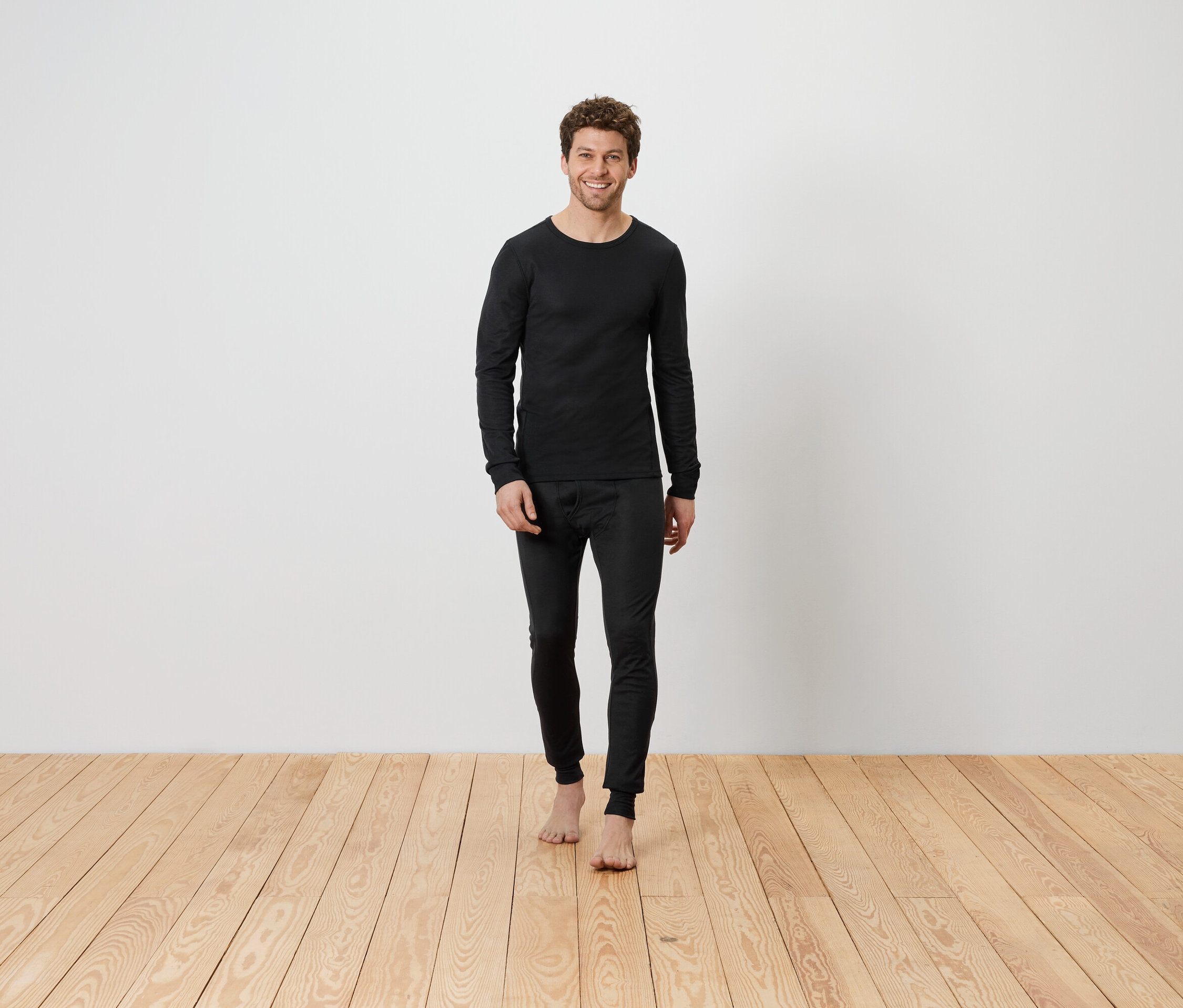 Férfi fekete termo alsóruházatban áll mezítláb a fapadlón egy fehér fal előtt.