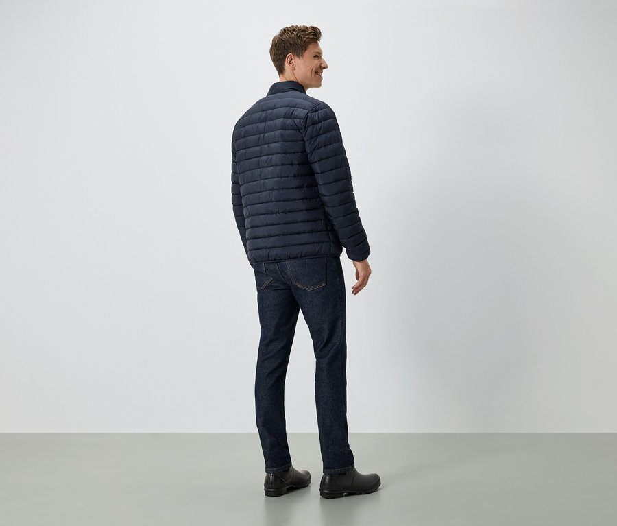 Egy férfi áll háttal a kamerának, sötétkék pehelykabátot, farmert és fekete cipőt visel.