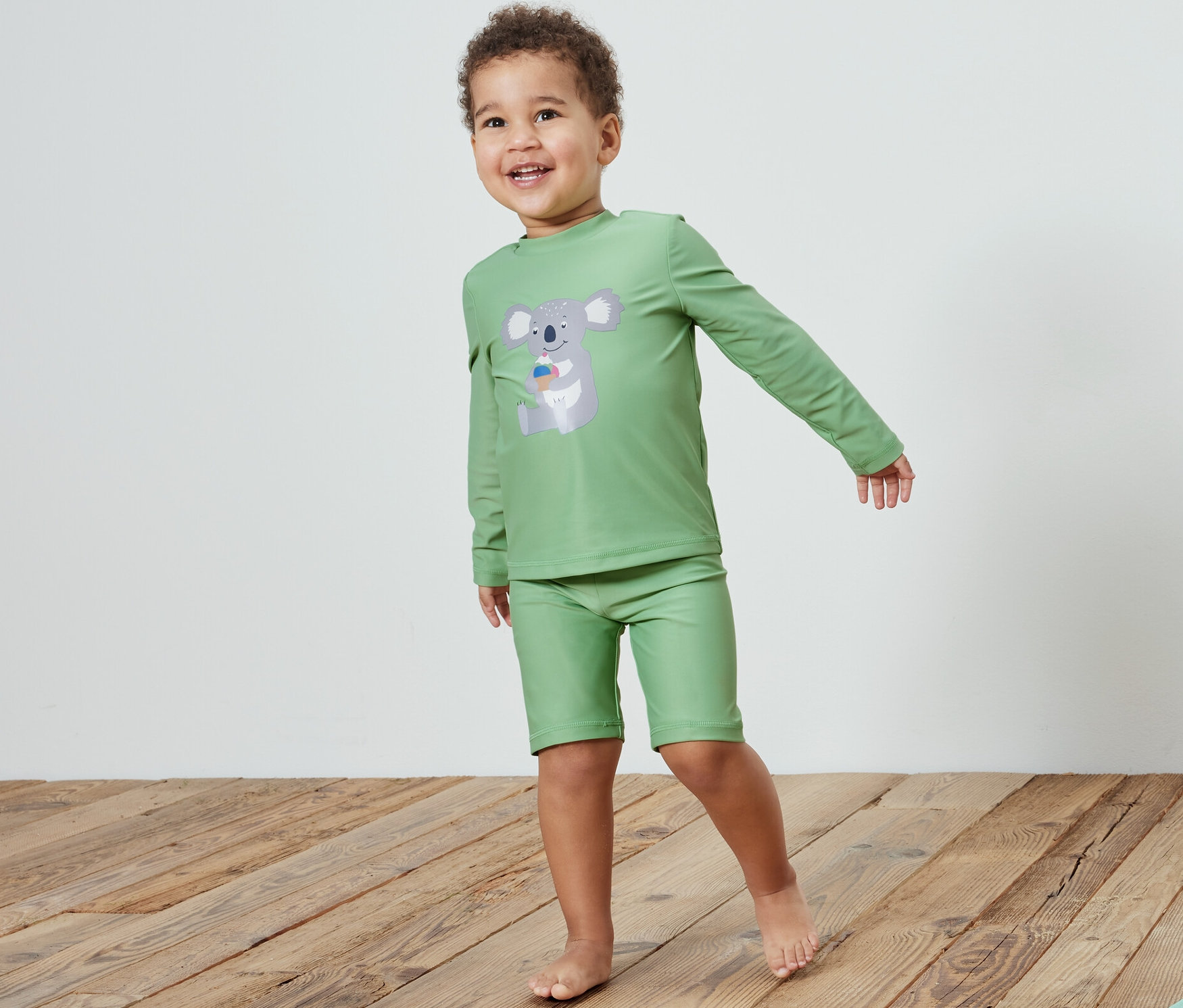 Egy fiú gyerek együttesben, 80-as fényvédő faktorral, koala, egy fapadlón áll egy játék mellett.