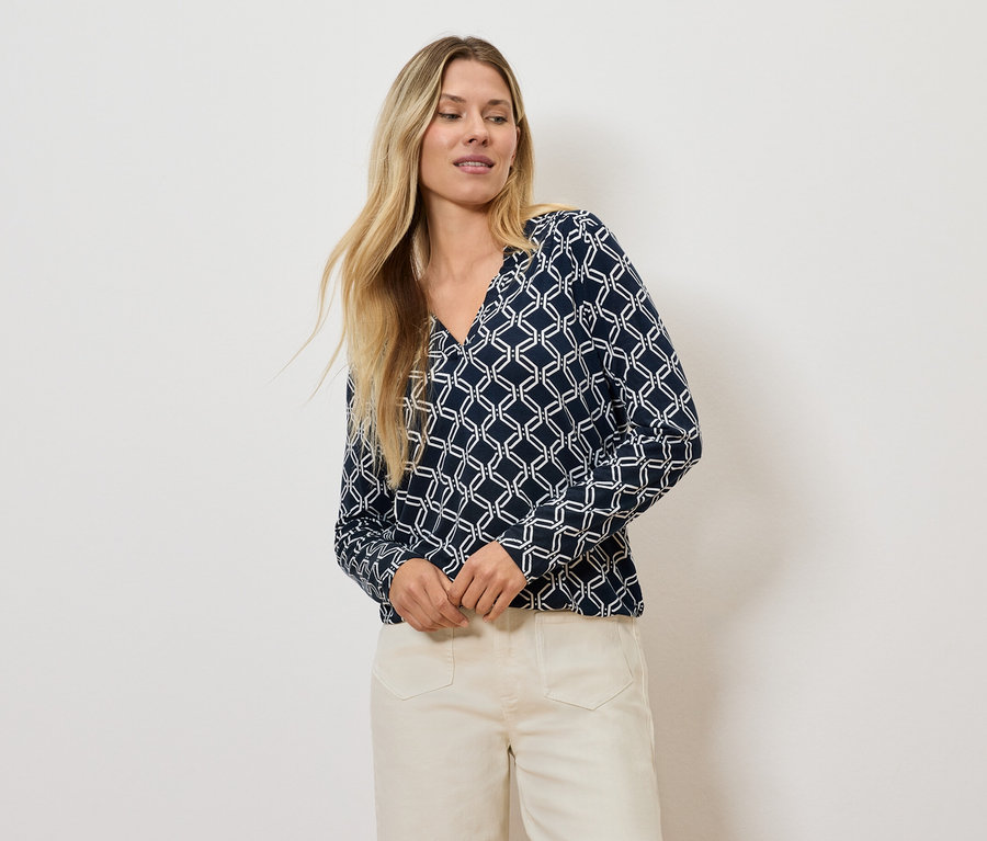 Szőke nő geometrikus mintás blúzban és világos nadrágban pózol.