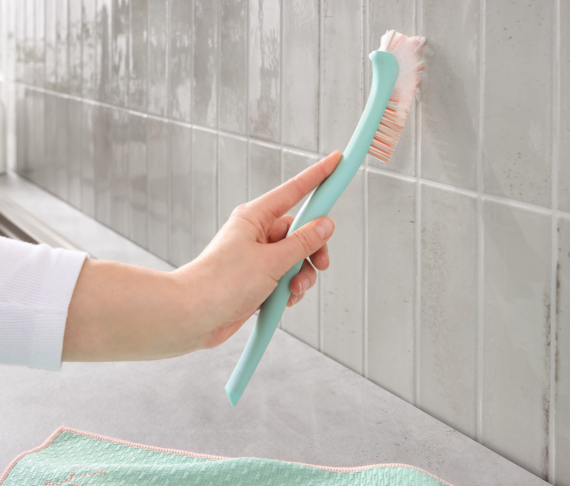 Egy kéz fugatisztító kefével tisztítja a piszkos fugákat a csempéken.