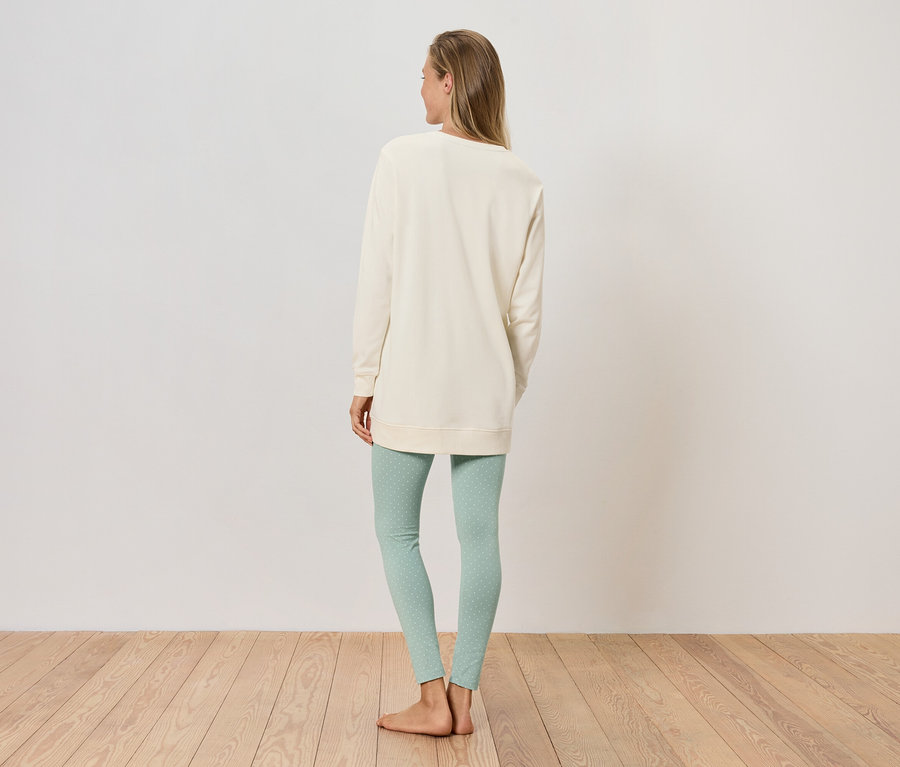 Nő áll háttal a kamerának, krémszínű pulóvert és világoszöld leggingset visel.