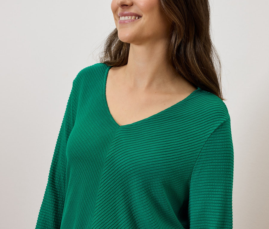 Mosolygó, barna hajú nő zöld, V-nyakú pulóvert visel.