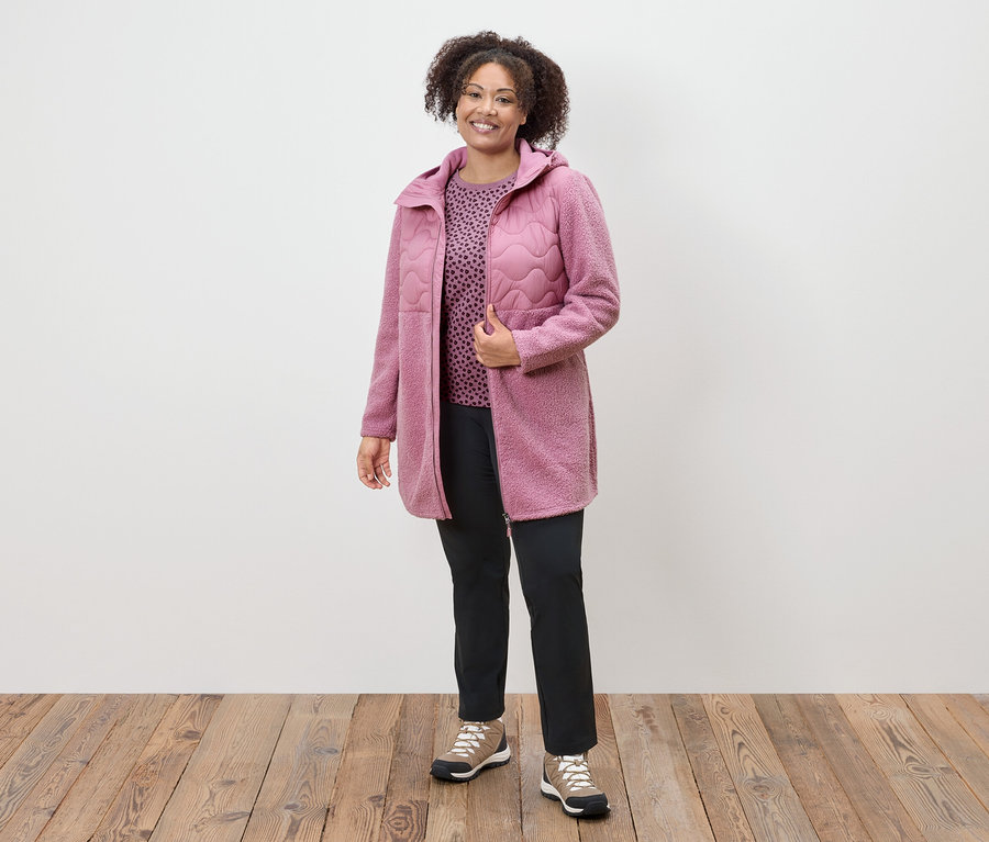 Egy nő rózsaszín kabátban, mintás felsőben és fekete nadrágban áll egy fapadlón.