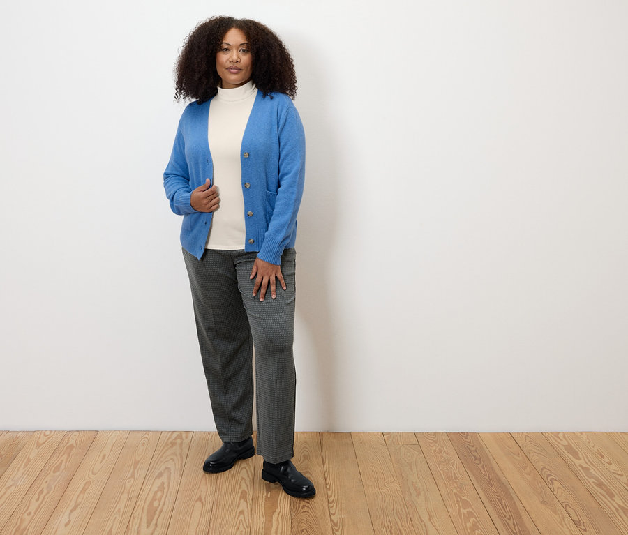 Nő áll a fapadlón, kockás, kényelmes jersey nadrágot, fehér garbót és kék kardigánt visel.