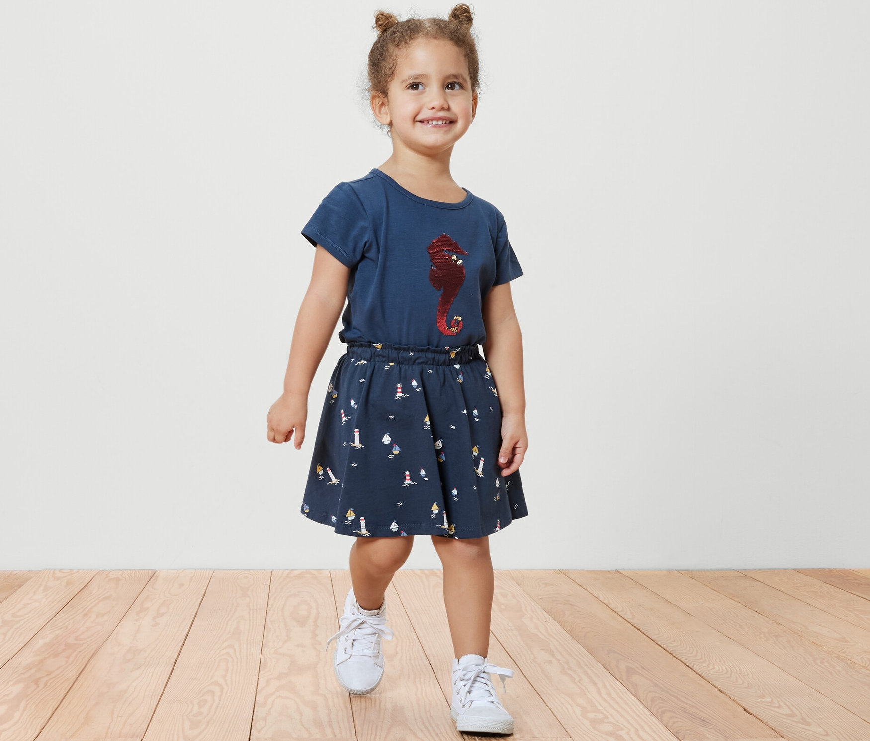 Mosolygó kislány sétál a fapadlón, kislány jerseyszoknyát visel nadrággal, tengerész stílusú, valamint egy kék pólót tengericsikó mintával.