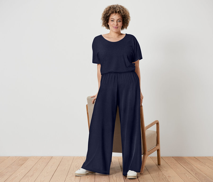 A göndör hajú nő sötétkék overallt és fehér cipőt visel, egy fa keretes szék mellett áll.