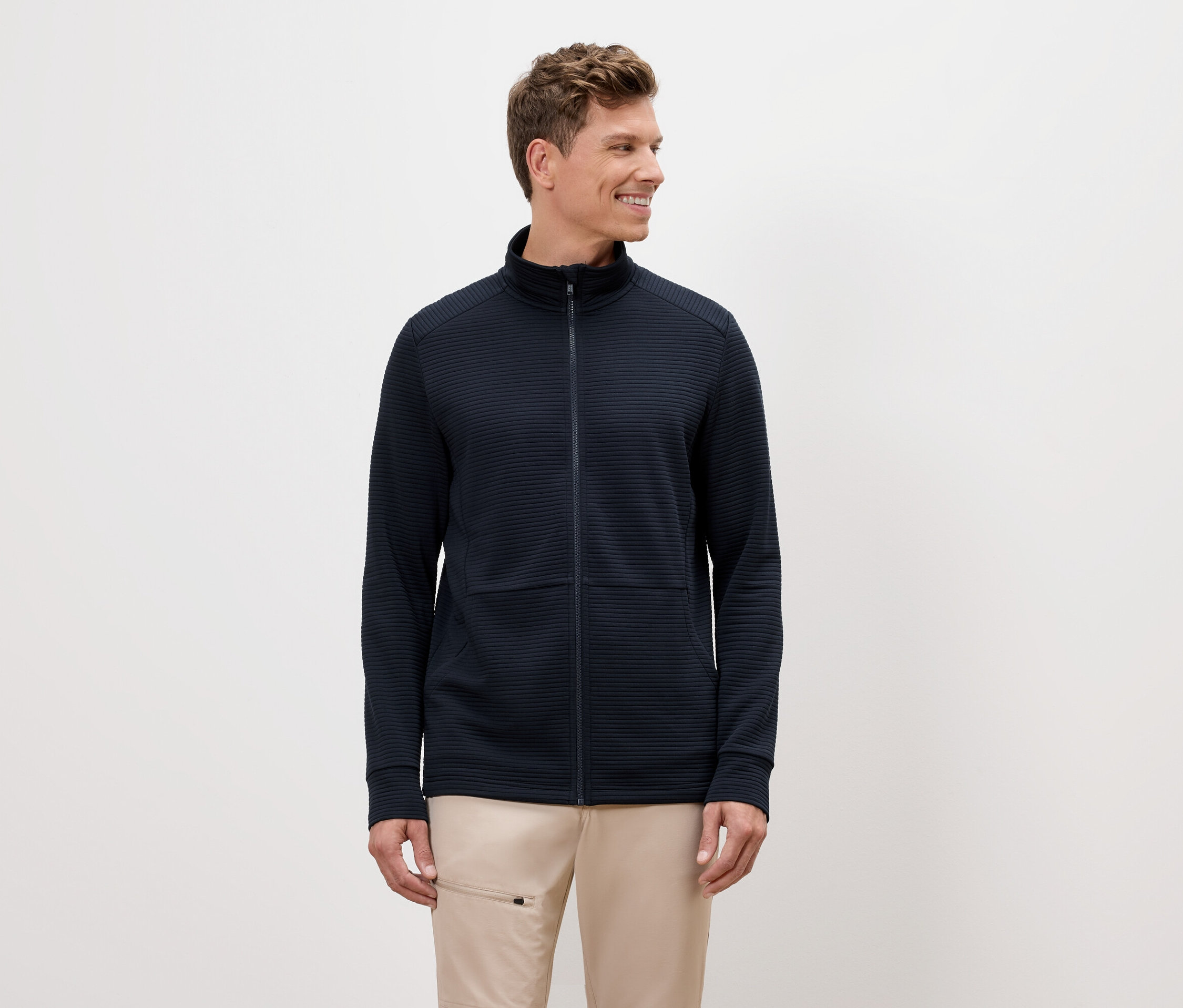 Egy szőke hajú férfi tengerészkék cipzáras kabátot és bézs nadrágot visel.