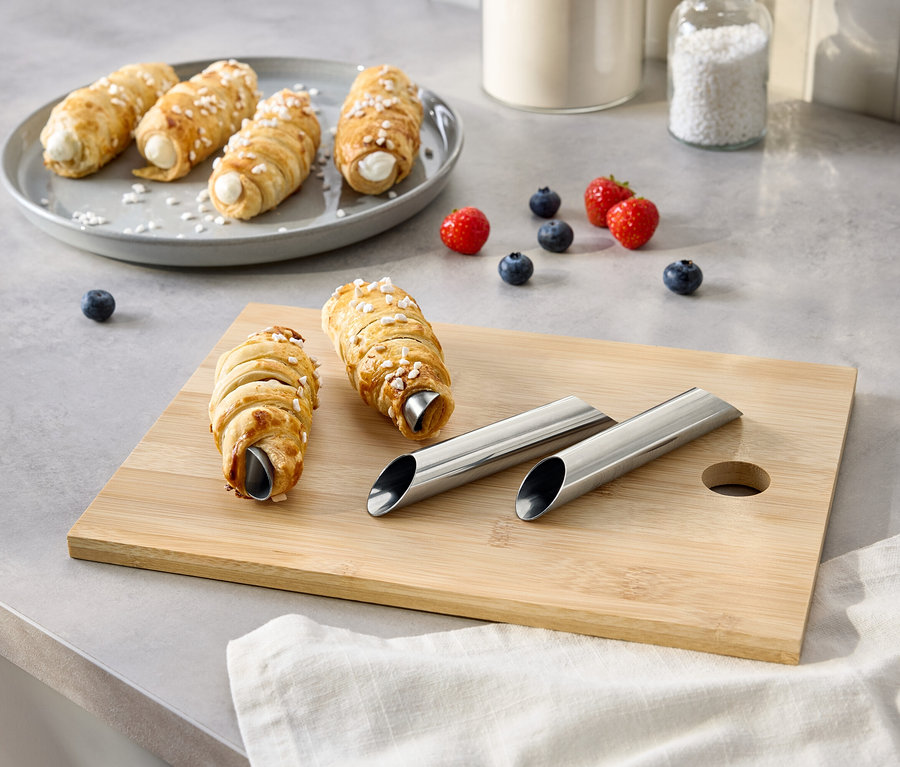 Az asztalon tejszínes tekercsek találhatók egy tányéron és egy vágódeszkán, bogyókkal, cukorral és fémformákkal együtt.