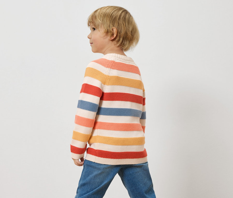 Egy szőke hajú gyermek csíkos pulóvert és farmert visel fehér háttér előtt.