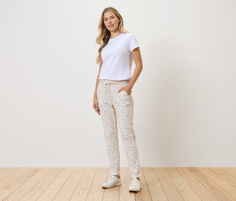 Nő áll a fal előtt, fehér pólót, mintás nadrágot és fehér tornacipőt visel.